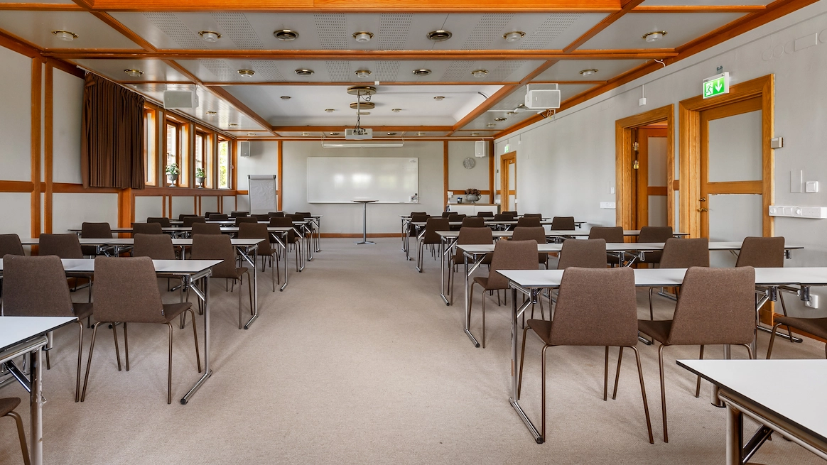 En bild av konferenslokalen Stora Tuna på Best Western Gustaf Wasa Hotel i Borlänge. Flera avlånga bord uppställda i två rader med fyra stolar bakom vardera. En whiteboardtavla och projektor syns i bakgrunden.