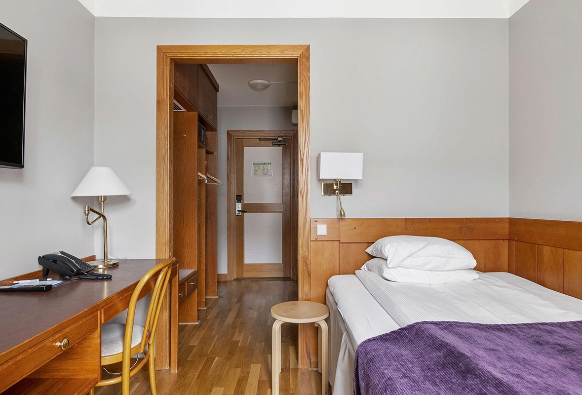 Ett enkelrum med en säng, ett skrivbord och en tv på väggen.