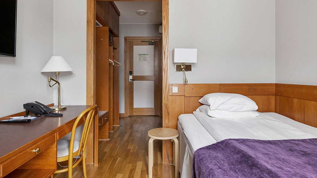 Ett enkelrum med en säng, ett skrivbord och en tv på väggen.