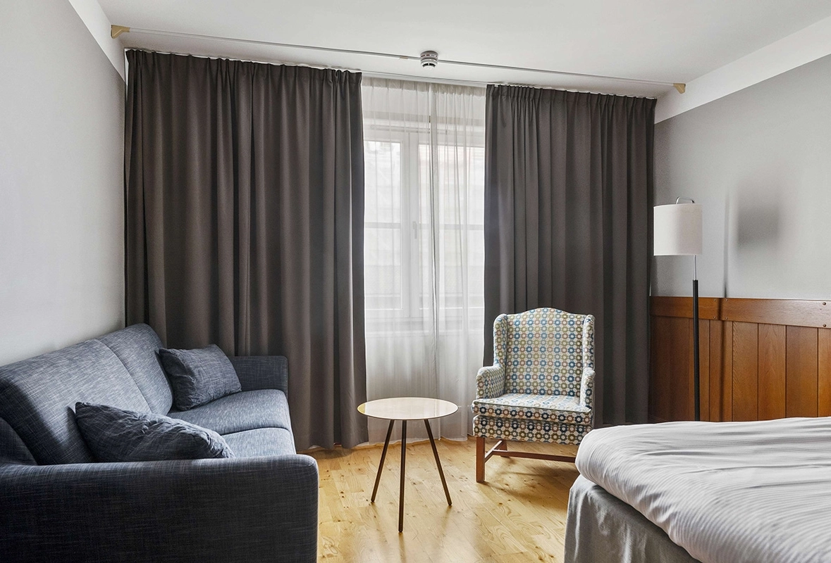 En soffa, ett litet bord, en fåtölj och ena hörnet av en stor säng. I bakgrunden ett par fönster.