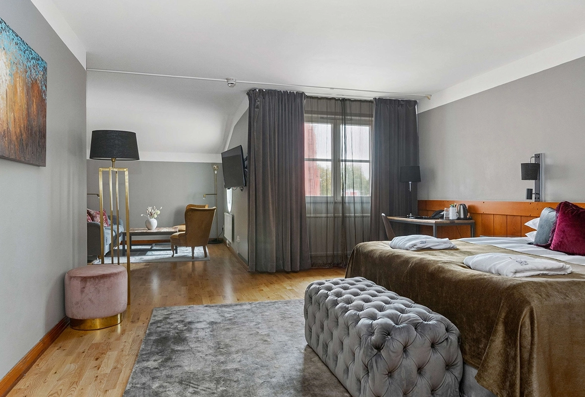 I bakgrunden syns en soffa och två fåtöljer runt ett lågt bord. I förgrunden en lyxig säng med en sittdyna vid fotändan. Ett skrivbord vid ett fönster.