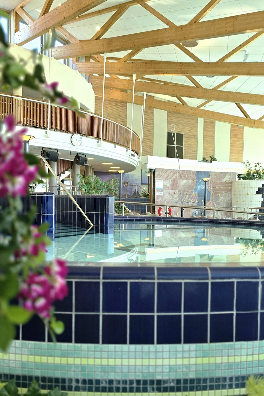 Inomhuspool med blå mosaik, omgiven av växter och träbjälkar i taket i ett ljust badhus