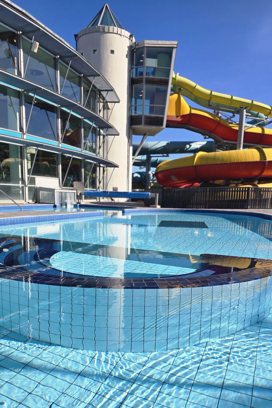En utomhuspool i soligt väder, och en rutschkana som sitter samman med ett stort torn i bakgrunden.