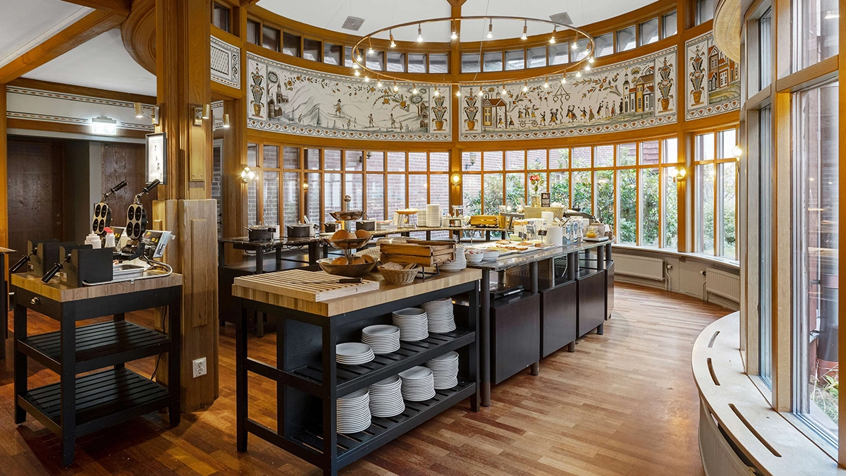 Frukostmatsalen på Gustaf Wasa Hotell. Ett stort rum med serveringsbord. I taket hänger en modern takkrona.