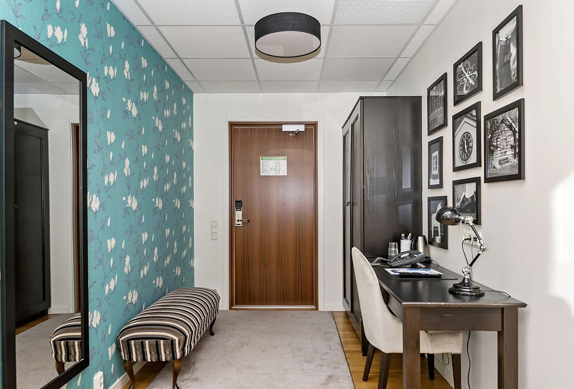 En rymlig hall med spegel, skrivbord med stol, samt en bänk. Ett antal inramade foton hänger på väggen ovanför skrivbordet.